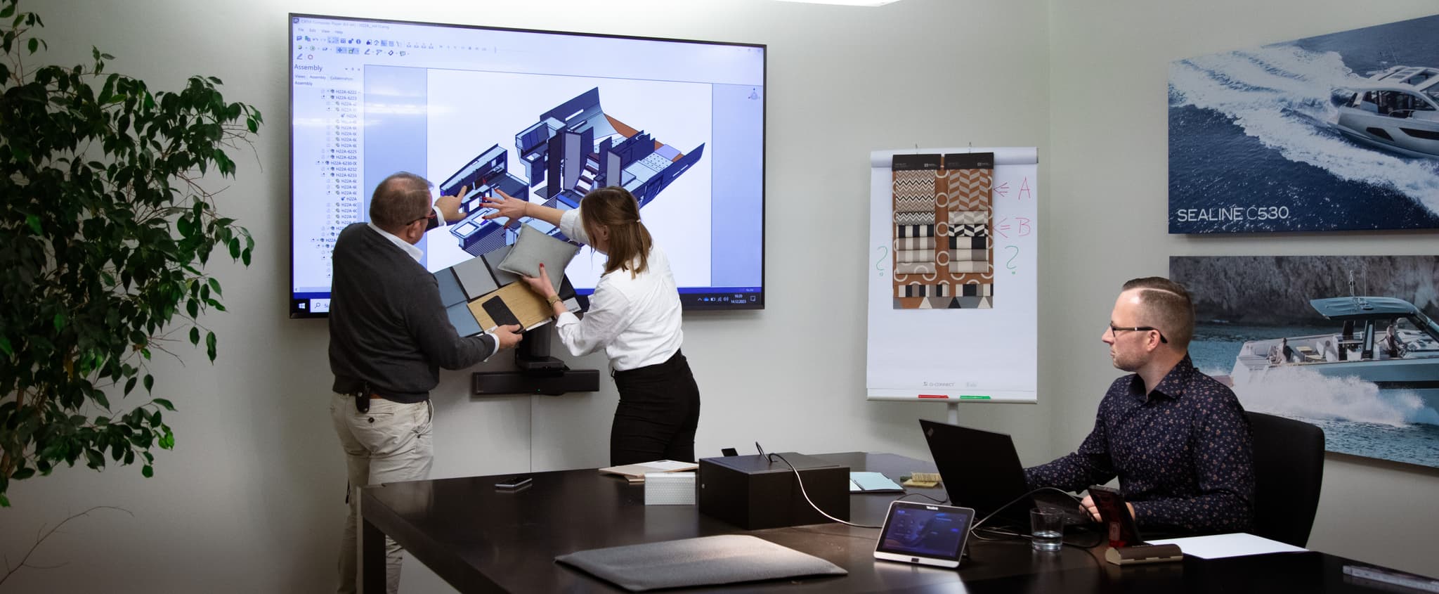 Professional team collaborating in a yacht design office with a 3D boat model on a large screen, material samples on a flipchart, and yacht posters on the wall, depicting a focused work environment.